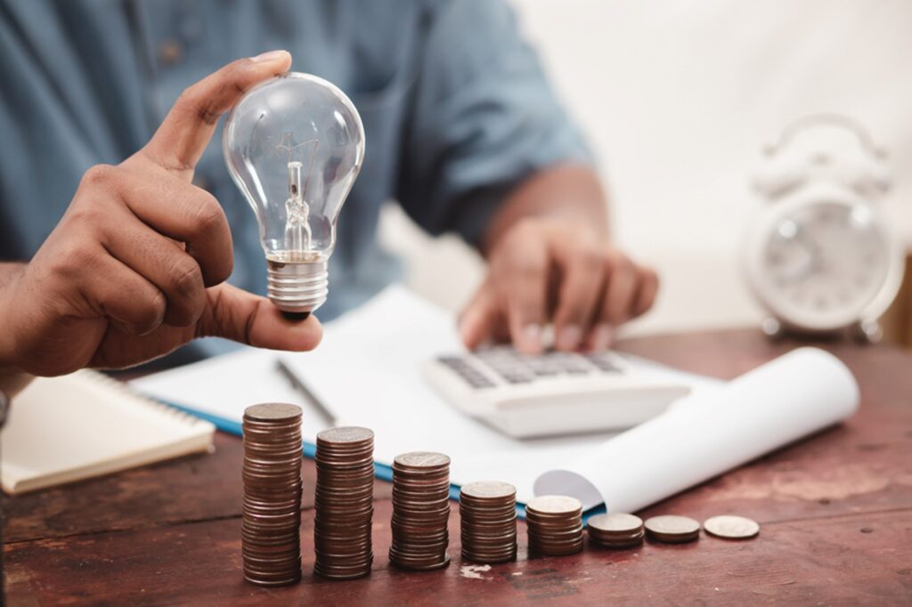 Des piles de monnaies, une ampoule dans la main d'un homme qui est en train de calculer les gains réalisés grâce aux travaux préconisés dans  l'audit énergétique. 