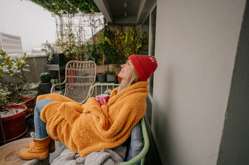 Une jeune femme profite de sa terrasse joliment aménagée en automne. Elle est assise sur un fauteuil, enroulée dans un plaid et avec une tasse de thé à la main. 