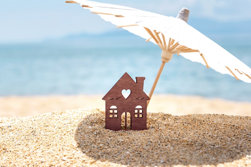 Maison et parapluie miniatures sur la plage, la mer bleue et le ciel sur le fond brouillé. Concept de projets immobiliers en été.