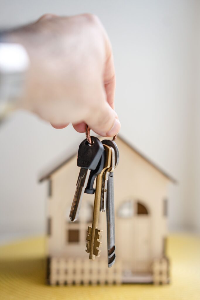 Une main tenant un trousseau de clé devant une maquette de maison en arrière-plan.